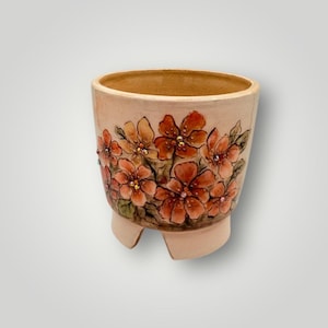 May include: A beige ceramic planter with a floral design. The flowers are painted in shades of orange and red with green leaves and small crystal accents. The planter has a rounded shape and three small feet.