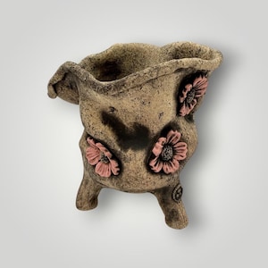 May include: A handcrafted, beige-colored ceramic planter with a textured surface. The planter features three pink flower embellishments and rests on three small legs. The unique design adds a rustic touch to any home decor.