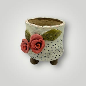 May include: A ceramic planter with a cream-colored base, adorned with black polka dots and two red rose accents. The planter has three small brown feet and green leaf details. A decorative item for plants.