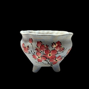 May include: A small, three-legged ceramic pot with a light blue exterior. The pot is decorated with red floral designs and black stems. The rim is a lighter shade of blue. The pot is set against a black background.