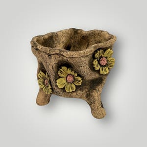 May include: A brown, three-legged planter with a textured surface. The planter is adorned with three yellow and pink flower accents. The planter has a rustic, weathered appearance, suitable for small plants or succulents.