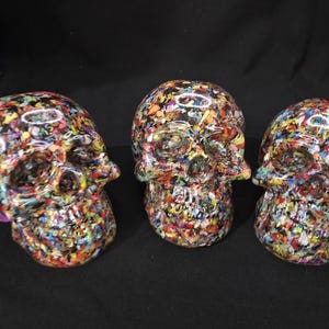 May include: Three skull-shaped decorative objects featuring a glossy finish and a dense, multi-colored confetti-like pattern composed of small, vibrant flecks of red, blue, yellow, green, and white embedded within a clear, smooth resin material.