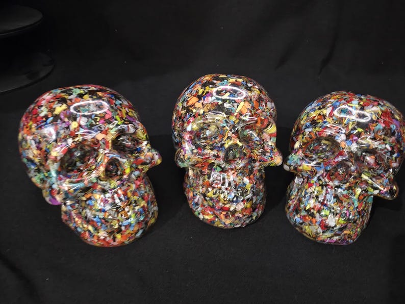 May include: Three skull-shaped decorative objects featuring a glossy finish and a dense, multi-colored confetti-like pattern composed of small, vibrant flecks of red, blue, yellow, green, and white embedded within a clear, smooth resin material.