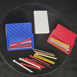 Op de afbeelding: Een bovenaanzicht toont drie kleine, rechthoekige containers en een verzameling kleurrijke haarspelden. Eén container is blauw, een andere rood en de derde wit. De haarspelden zijn in verschillende kleuren, waaronder rood, wit, zwart en geel.