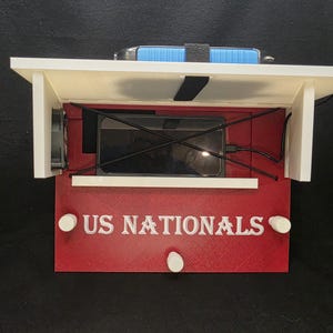 Op de afbeelding: Een houten oplaadstation met een rood voorpaneel en witte accenten. Op het voorpaneel staat de tekst "US NATIONALS" in het wit. Binnenin is een zwarte smartphone te zien, met oplaadkabels. Bovenop ligt een blauw object.