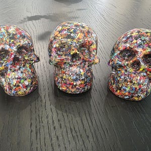 May include: Three decorative skull figurines featuring a dense, multi-colored confetti-like pattern throughout. Each skull displays a glossy finish with a vibrant mix of red, blue, yellow, green, and black fragments embedded across the entire surface.