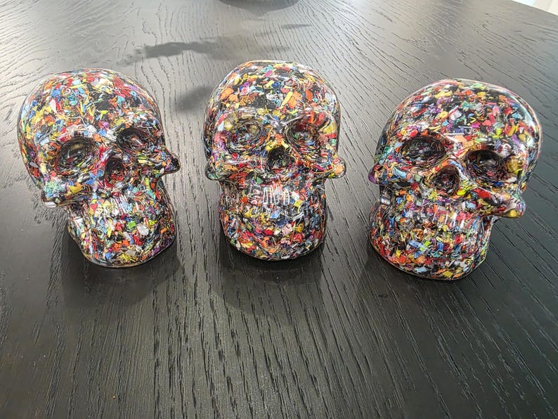 May include: Three decorative skull figurines featuring a dense, multi-colored confetti-like pattern throughout. Each skull displays a glossy finish with a vibrant mix of red, blue, yellow, green, and black fragments embedded across the entire surface.