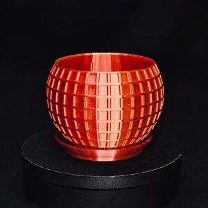 May include: A spherical orange planter with a grid-like textured surface and a matching circular base tray. The pot features a repeating square pattern across its exterior, creating a geometric design that wraps around the entire rounded body.