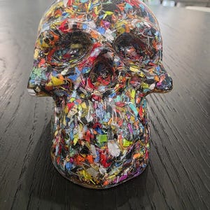 May include: A skull-shaped sculpture made of clear resin embedded with a dense, multi-colored mosaic of small, fragmented plastic pieces in shades of red, blue, yellow, green, and black, creating a textured, confetti-like appearance throughout.