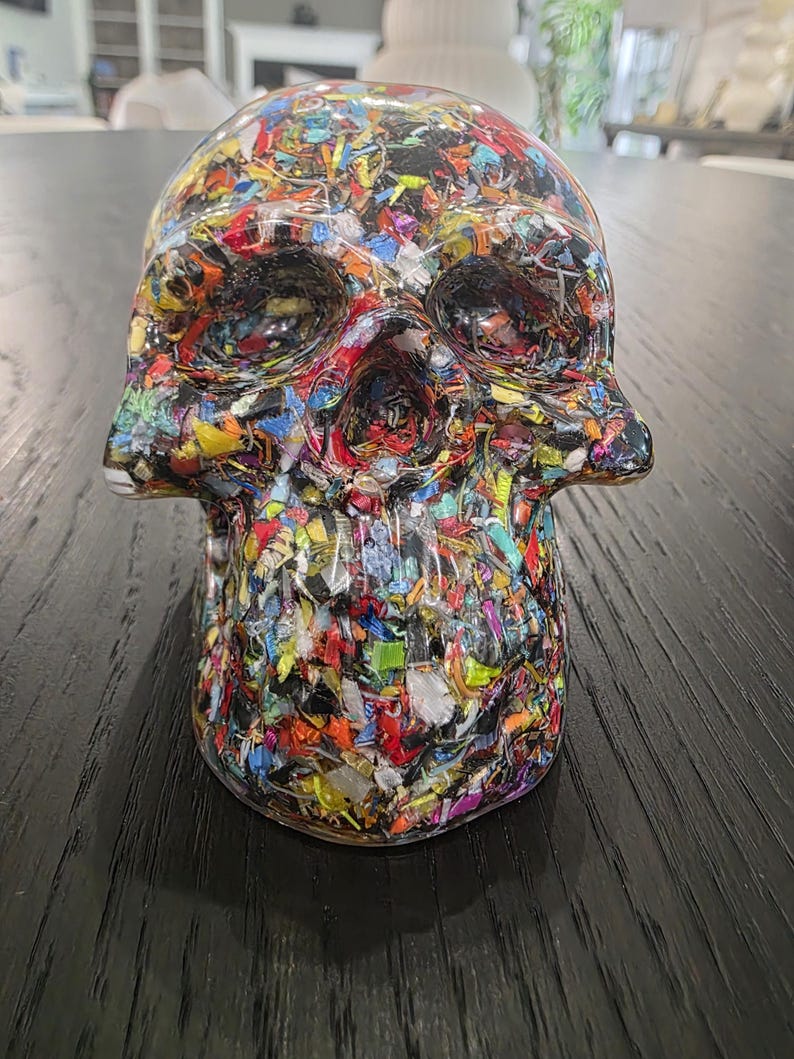 May include: A skull-shaped sculpture made of clear resin embedded with a dense, multi-colored mosaic of small, fragmented plastic pieces in shades of red, blue, yellow, green, and black, creating a textured, confetti-like appearance throughout.