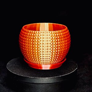 May include: A spherical orange planter featuring a repeating geometric pyramid texture across its exterior surface. The planter includes a matching circular base tray and has a smooth, flat rim at the top opening, showcasing a consistent 3D printed finish.