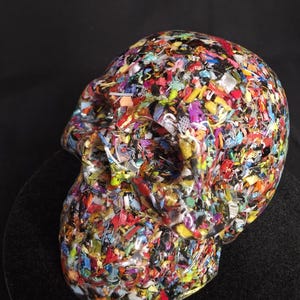 May include: A human skull sculpture crafted from a clear resin base embedded with a dense, multi-colored mosaic of tiny plastic fragments, including red, blue, yellow, green, and black pieces, creating a textured, confetti-like appearance.