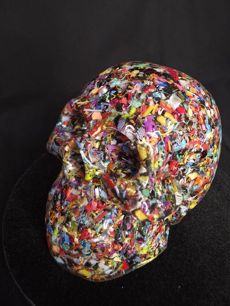 May include: A human skull sculpture crafted from a clear resin base embedded with a dense, multi-colored mosaic of tiny plastic fragments, including red, blue, yellow, green, and black pieces, creating a textured, confetti-like appearance.