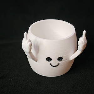 May include: A white, cylindrical 3D-printed planter featuring a small, smiling face with black eyes and a mouth. The planter has two arms extending from its sides, each with a hand raised, displaying a middle finger gesture in a defiant pose.