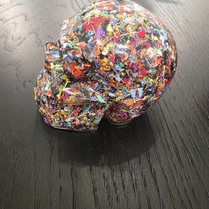 May include: A human skull sculpture made of clear resin embedded with a dense mixture of multicolored plastic fragments. The skull features a smooth, glossy finish that encases the vibrant, confetti-like pieces of red, blue, yellow, and green.