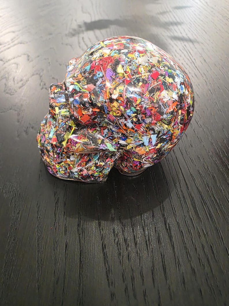 May include: A human skull sculpture made of clear resin embedded with a dense mixture of multicolored plastic fragments. The skull features a smooth, glossy finish that encases the vibrant, confetti-like pieces of red, blue, yellow, and green.