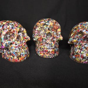 May include: Three skull-shaped decorative objects featuring a dense, multi-colored confetti-like pattern. The surface of each skull is covered in small, irregular flecks of red, blue, yellow, green, and black, creating a vibrant, textured appearance.