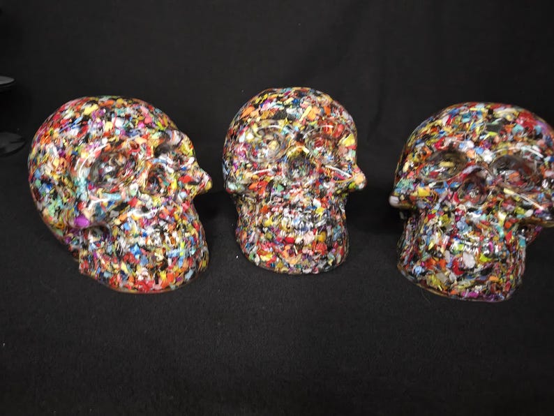May include: Three skull-shaped decorative objects featuring a dense, multi-colored confetti-like pattern. The surface of each skull is covered in small, irregular flecks of red, blue, yellow, green, and black, creating a vibrant, textured appearance.