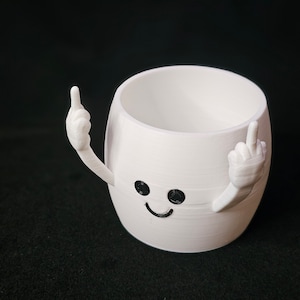 May include: A white cylindrical planter with a smooth finish, featuring a small black smiling face and two arms raised on either side, each with a finger extended in a gesture. The object has a hollow interior and a flat base for stability.
