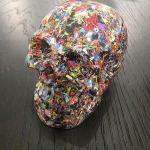 May include: A human skull sculpture crafted from a clear resin base embedded with a dense, multi-colored mosaic of small, fragmented plastic pieces in shades of red, blue, yellow, green, and purple, featuring a smooth, glossy, reflective finish.