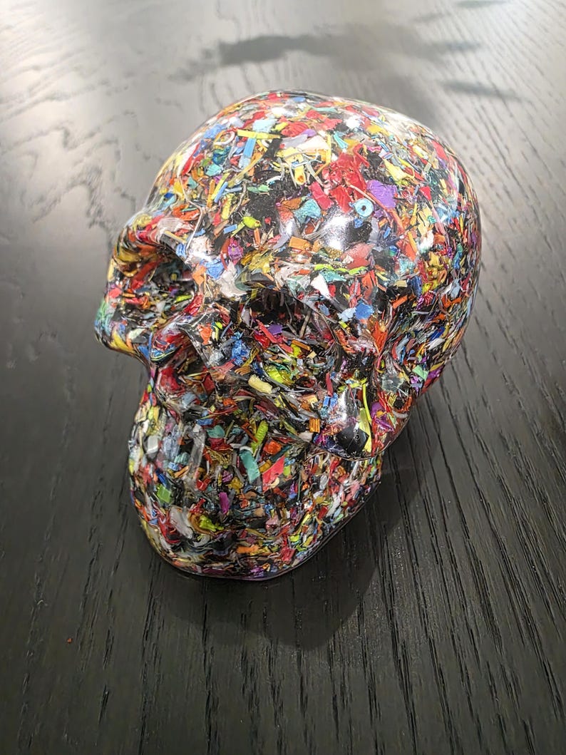 May include: A human skull sculpture crafted from a clear resin base embedded with a dense, multi-colored mosaic of small, fragmented plastic pieces in shades of red, blue, yellow, green, and purple, featuring a smooth, glossy, reflective finish.