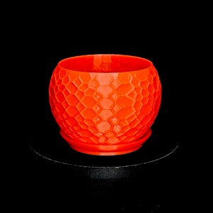 May include: A bright orange, spherical planter featuring a textured, geometric honeycomb pattern across its exterior surface. The planter includes a matching circular base tray and has a wide, open top, showcasing a consistent, vibrant colour throughout.