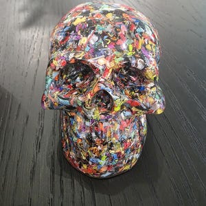 May include: A human skull sculpture crafted from a dense mosaic of multicolored plastic fragments. The surface features a chaotic blend of red, blue, yellow, green, and black flecks embedded within a clear, glossy resin material.