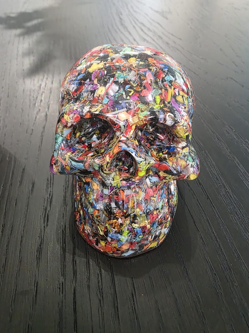 May include: A human skull sculpture crafted from a dense mosaic of multicolored plastic fragments. The surface features a chaotic blend of red, blue, yellow, green, and black flecks embedded within a clear, glossy resin material.