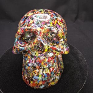 May include: A human skull sculpture crafted from clear resin embedded with a dense mixture of multicolored plastic confetti, including red, yellow, blue, green, and purple fragments, creating a vibrant, textured, and translucent appearance.
