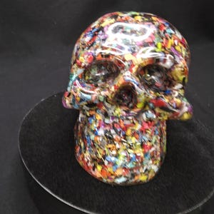May include: A skull sculpture featuring a dense, multi-colored mosaic pattern composed of tiny, fragmented pieces in shades of red, blue, yellow, green, and white. The surface has a smooth, glossy finish that reflects light across the entire form.