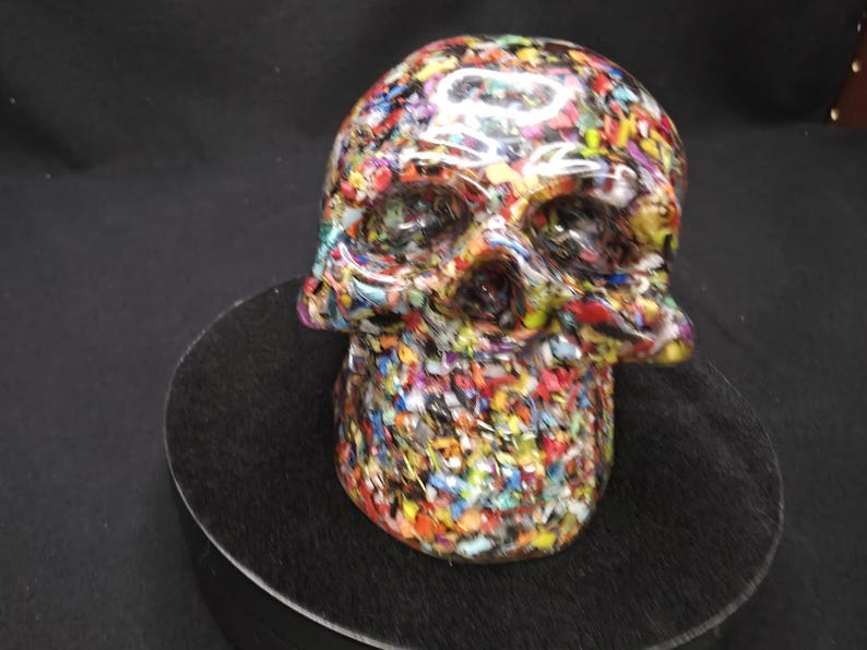 May include: A skull sculpture featuring a dense, multi-colored mosaic pattern composed of tiny, fragmented pieces in shades of red, blue, yellow, green, and white. The surface has a smooth, glossy finish that reflects light across the entire form.