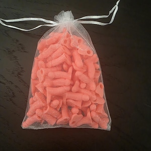 Puede incluir: Una bolsa de organza blanca transparente llena de numerosos caramelos de goma rosas. Los caramelos tienen forma fálica y están ligeramente espolvoreados. La bolsa está atada con una cinta blanca.
