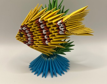 May include: A yellow origami fish with red and white stripes, standing on a blue origami base with green origami leaves behind it.