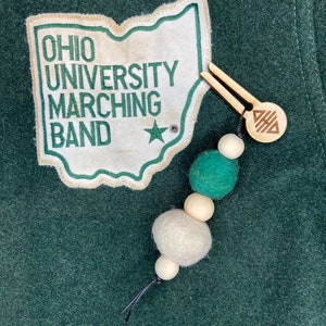 Op de afbeelding: Een groene viltpatch met de tekst "OHIO UNIVERSITY MARCHING BAND" en een witte ster. Een houten hanger met een geometrisch ontwerp is bevestigd aan een touwtje met groene en witte viltballen.