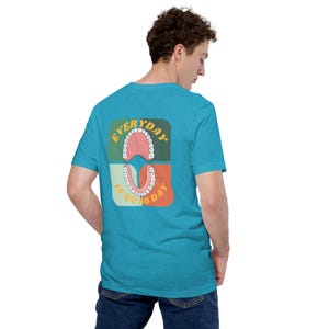 Puede incluir: Una camiseta turquesa con un diseño gráfico en la espalda. El diseño presenta dos arcos dentales estilizados, uno encima del otro, con el texto "EVERYDAY IS QUAD DAY" en una fuente retro. La camiseta está hecha de un material suave.
