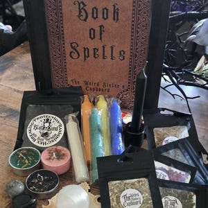 May include: A collection of mystical items, including a book titled "Book of Spells", candles in various colors, small bags of herbs, and crystals. The items are arranged on a wooden surface, creating a witchy aesthetic.
