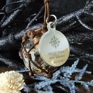 May include: A brown cord necklace with a glass bottle pendant containing a compass and a cork stopper. The pendant is attached to a round silver tag with the inscription "not all those who wander are lost" and a compass design.