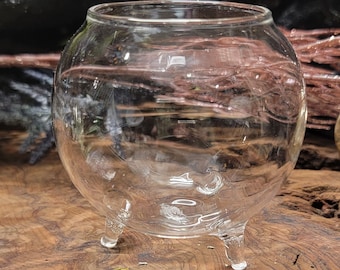 May include: A clear glass terrarium with three legs. The terrarium is round and has a wide opening at the top.