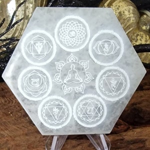 May include: White hexagonal selenite charging plate with seven etched chakra symbols, including the crown, third eye, throat, heart, solar plexus, sacral, and root chakras. The plate is on a wooden stand.