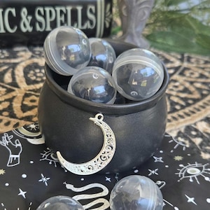 May include: A black cauldron filled with several clear, spherical plastic capsules. A silver-toned metal crescent moon charm with intricate cut-out patterns hangs from the rim of the cauldron. Two additional clear capsules rest in front.