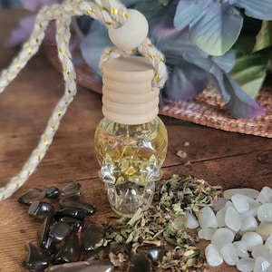 May include: A glass bottle shaped like a skull filled with a yellow liquid and a wooden stopper. The bottle is hanging from a white cord with gold accents. The bottle is surrounded by small black and white stones and dried herbs.