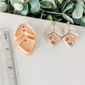 May include: Two pairs of handmade earrings with a white and gray marbled design. The earrings are decorated with small pink and gold flowers. A ruler is in the background.