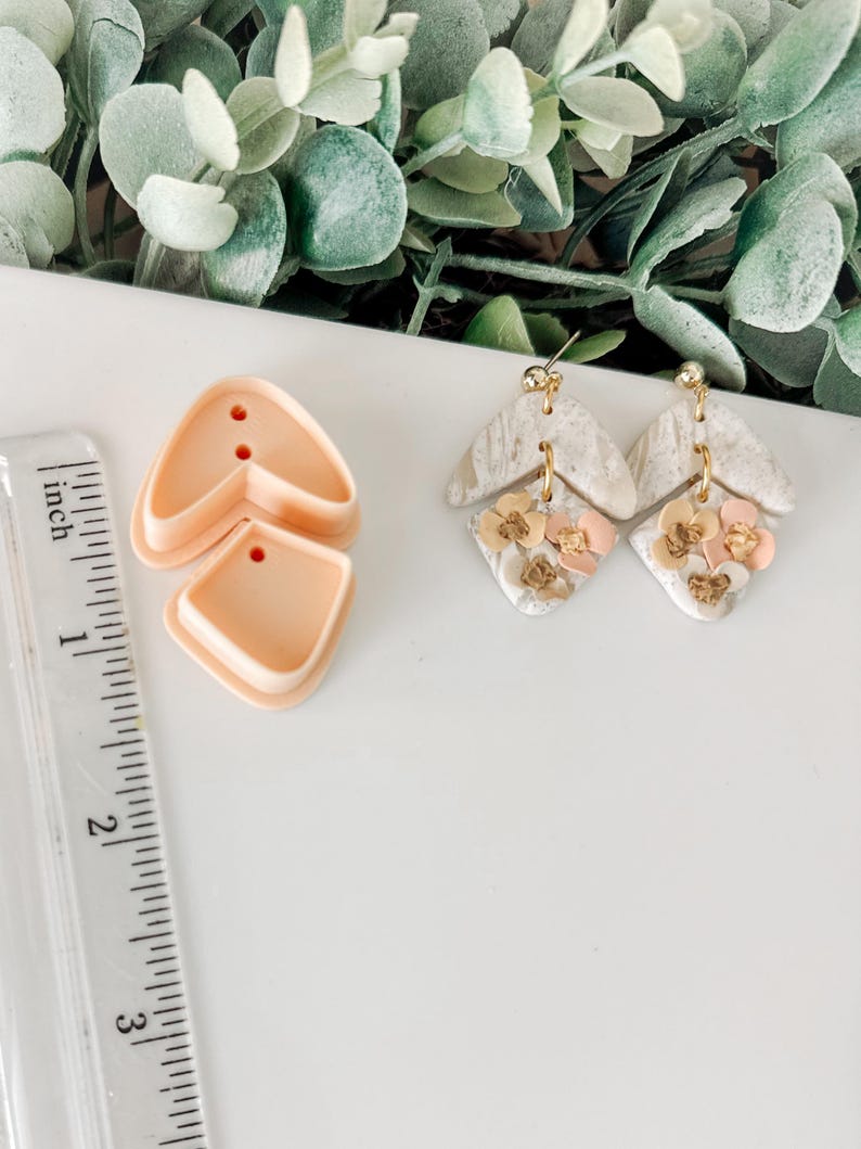 May include: Two pairs of handmade earrings with a white and gray marbled design. The earrings are decorated with small pink and gold flowers. A ruler is in the background.