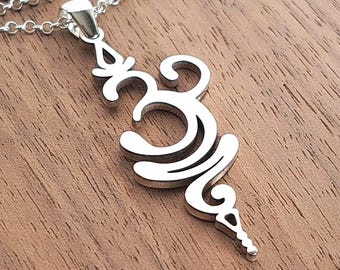 May include: A silver-colored pendant featuring an elongated, stylized Om symbol with smooth, curved lines. The pendant is attached to a delicate, fine-link silver chain that loops across the frame, showcasing a polished, reflective metallic finish.