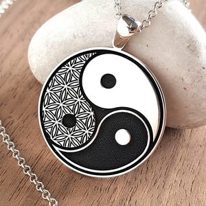 Könnte beinhalten: Eine silberfarbene, kreisförmige Anhängerkette mit einem schwarz-weißen Yin-Yang-Symbol. Eine Hälfte des Yin-Yang zeigt ein detailliertes geometrisches Muster der Blume des Lebens. Der Anhänger hängt an einer feinen, silberfarbenen Metallkette.