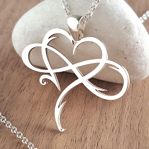 May include: A silver-coloured necklace featuring a pendant that combines an infinity symbol with a heart shape. The pendant is suspended from a delicate silver-coloured chain with a lobster claw clasp. The design is polished and reflective in appearance.