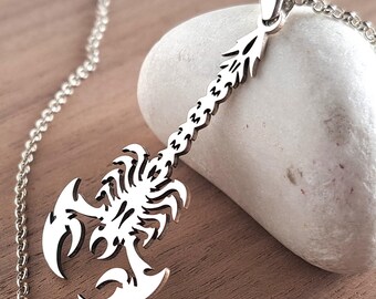 May include: A silver pendant necklace with a scorpion design. The pendant is detailed and hangs from a fine chain. The necklace is displayed on a white stone and a wooden surface.