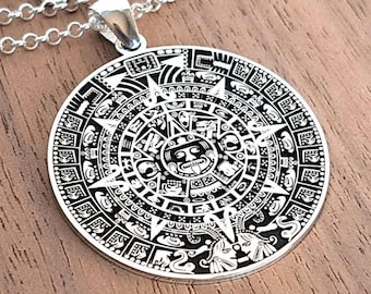 Puede incluir: Un colgante circular de color plateado con un diseño detallado de calendario azteca grabado en negro, suspendido de una cadena de eslabones plateada a juego. El colgante muestra intrincados patrones geométricos y un motivo central de rostro.