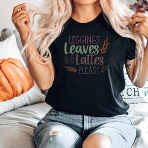 May include: A black t-shirt with the text "leggings leaves & lattes please" in a fall-themed font with brown, green, and pink accents.