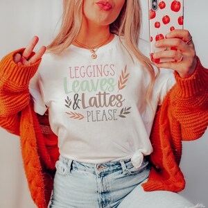 May include: A white t-shirt with the text "leggings leaves & lattes please" in a fall-themed font with brown and green leaves.